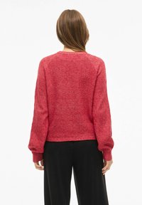 Punane eeritav kampsun tekstuurse mustriga, lahtine lõige, pikad varrukad ja ümmargune kaelus; koos mustade pükstega.