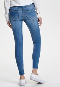 Jeans ajustados en denim azul medio, con una cintura de altura media, dos bolsillos traseros y dobladillo deshilachado. Llevados con zapatillas blancas.