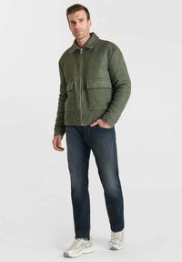 Chaqueta de mezcla de lana verde con cuello alto, cierre frontal y dos grandes bolsillos de parche, combinada con jeans oscuros y zapatillas claras.