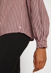 Blouse à manches longues à carreaux bordeaux et blancs avec des poignets à volants, portée avec un pantalon noir ajusté, vue sur le bras et le côté d'une personne.