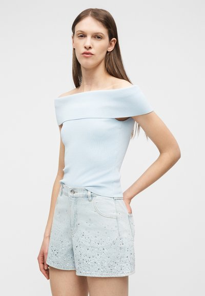 Γυναίκα με μακριά καστανά μαλλιά, που φοράει ανοιχτό γαλάζιο τοπ έξω από τον ώμο και ανοιχτά τζιν σορτς με διακοσμητικά στοιχεία, στέκεται με το ένα χέρι στη μέση.