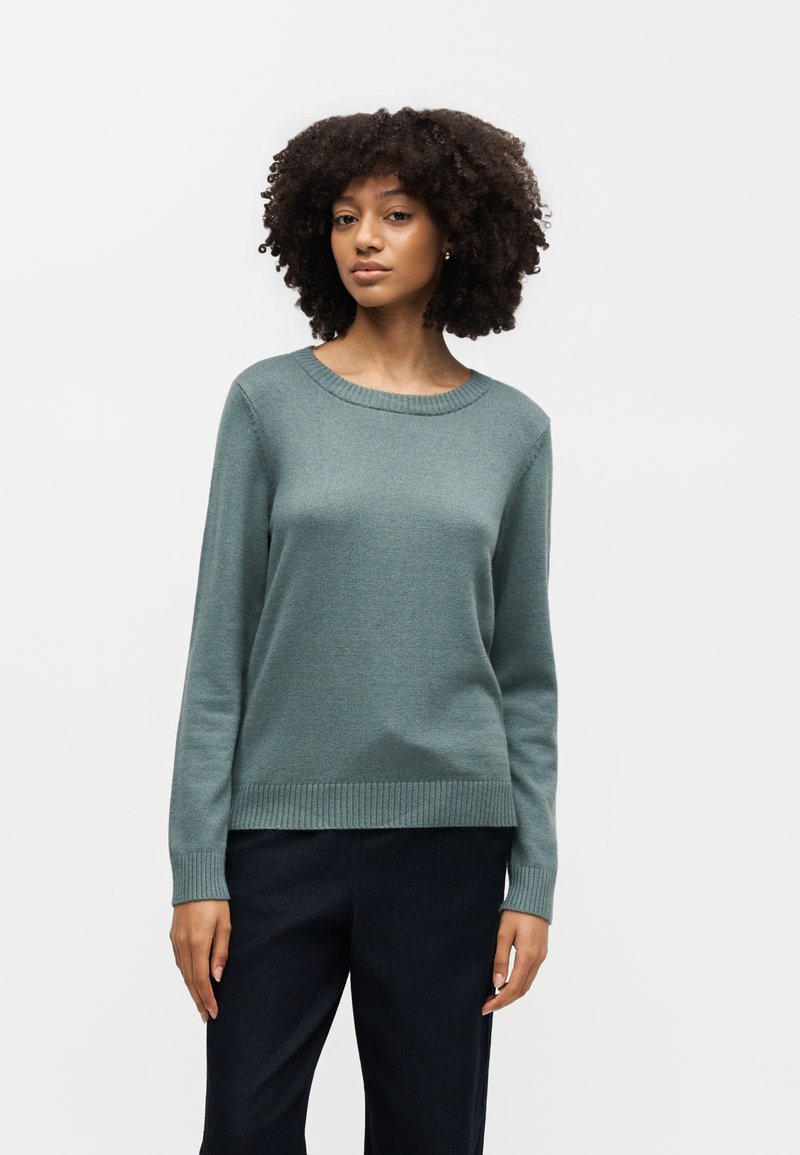Femme aux cheveux bouclés portant un pull vert doux à col rond et un pantalon noir, debout devant un fond clair uni.