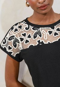 Top negro con tejido texturizado que presenta un yugo de encaje blanco adornado con patrones florales y de corazones. Cuello redondeado, mangas cortas.
