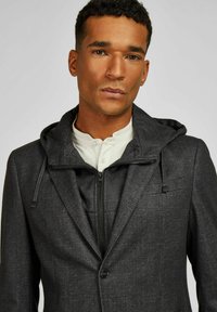 Donkergrijze gestructureerde blazer met een gevoerde capuchon, voorzien van een rits aan de voorkant en een knoopsluiting. Aangevuld met trekkoorden en een zijzak.