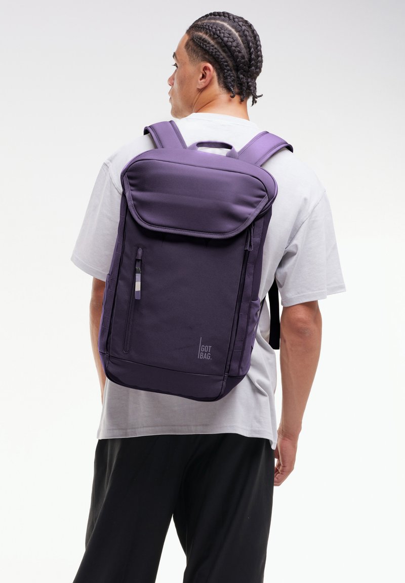 Homme aux cheveux tressés portant une chemise blanche et un pantalon noir, portant un grand sac à dos violet étiqueté "GOT BAG" sur un fond uni.
