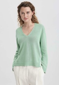 V-Ausschnitt, weicher Strickpullover in Hellgrün. Mit gerippten Bündchen und Saum, lockerer Schnitt mit seitlichen Schlitzen, kombiniert mit hellen Hosen.