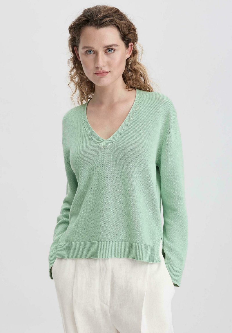 V-Ausschnitt, weicher Strickpullover in Hellgrün. Mit gerippten Bündchen und Saum, lockerer Schnitt mit seitlichen Schlitzen, kombiniert mit hellen Hosen.