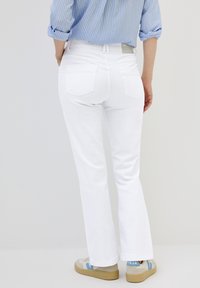 Vita utsvängda jeans med två bakfickor, i kombination med beige och blå sneakers; en randig blå skjorta är delvis synlig, med uppvikt ärm.