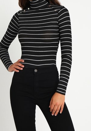 Femme portant un pull à col roulé rayé horizontal noir et blanc avec des détails de boutons et un pantalon noir ajusté, main sur la hanche.