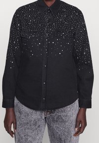 Black denim shirt with silver decorative studs, featuring two front pockets, a classic collar, and button closures. Textured fabric with a subtle sheen.