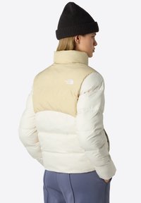 Puffjacka med en färgsättning i kräm och beige. Har en hög krage, horisontell quiltad struktur och en logo på baksidan.