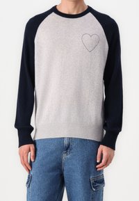 Homme portant un pull gris clair avec des manches bleu marine et un petit cœur brodé sur la poitrine, associé à un jean cargo bleu.