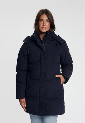 Vrouw met donker haar die een marineblauwe gewatteerde winterjas met capuchon draagt, staat met één hand in een zak tegen een effen achtergrond.