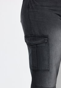 Pantaloni cargo neri in denim aderenti con tasca laterale, caratterizzati da una texture liscia e una leggera scoloritura, dettagli di cucitura visibili e un design strutturato.