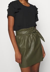 Schwarze Bluse mit Rüschensleeves, kombiniert mit einem hochgeschnittenen, olivgrünen Leder-Minirock mit einem gebundenen Gürtel. Glatte Texturen und strukturiertes Design.