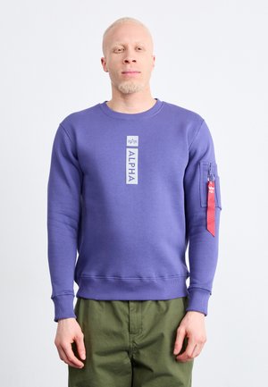 Mann trägt lila Sweatshirt mit dem Schriftzug "ALPHA" und Reißverschlusstasche mit rotem Etikett am Ärmel, kombiniert mit olivgrünen Hosen, vor weißem Hintergrund stehend.