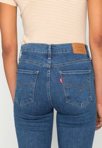 Jeans de ganga azuis com cintura alta, apresentando dois bolsos traseiros e uma etiqueta de couro castanho na cintura. Detalhes de costura ao redor dos bolsos.