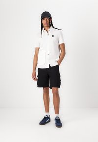 Hvit kortermet skjorte med frontlomme og svart knapp, parret med svarte cargo-shorts og marineblå sneakers med lyseblå striper.