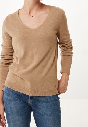 Woman wearing a beige V-neck sweater and blue jeans, with a delicate black pendant necklace, posing with one hand in pocket.