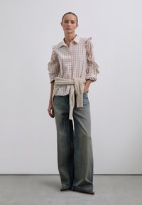 Camisa a cuadros rosa claro y blanca con mangas acampanadas, combinada con jeans de mezclilla deslavados de pierna ancha y un suéter beige claro atado a la cintura.