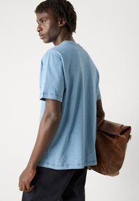 Camiseta de algodón azul claro con mangas cortas, cuello redondo y un corte relajado, combinada con un bolso de cuero marrón. Diseño sencillo y textura suave.