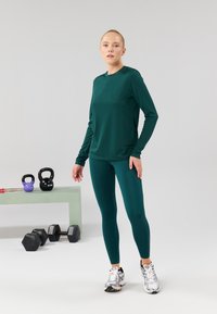 Grünes, langärmliges Sportoberteil mit passenden Leggings, aus glattem Stoff und in taillierter Passform, kombiniert mit silbernen Sportschuhen.