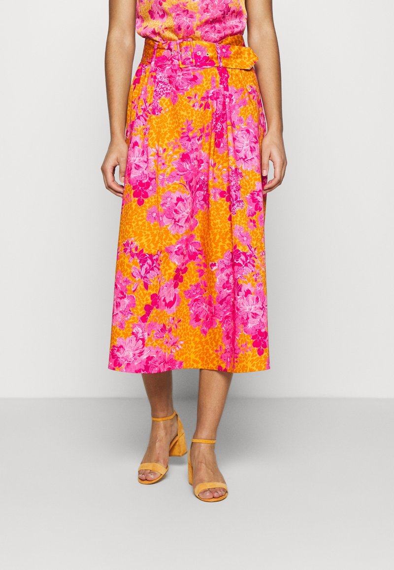 Femme portant une jupe florale éclatante avec des fleurs roses et orange, ceinture assortie, sandales jaunes à talons, debout devant un fond uni.