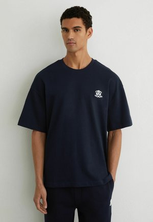 Ung mand iført en ensfarvet mørkeblå T-shirt med et lille hvidt logo på venstre bryst, stående mod en lysegrå væg.