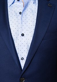 Granatowy, teksturowany żakiet założony na jasnoniebieską koszulę z małymi granatowymi kwadratowymi wzorami i błyszczącymi czarnymi guzikami. Zauważalny detal akcentujący klapę.