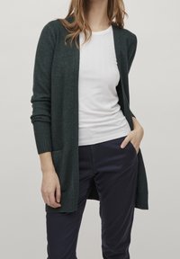 Cardigan verde oscuro hecho de un suave tejido de punto, con mangas largas, un frente abierto y dos bolsillos laterales, llevado sobre una camiseta blanca ajustada.