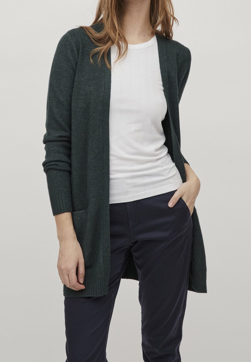 Cardigan verde oscuro hecho de un suave tejido de punto, con mangas largas, un frente abierto y dos bolsillos laterales, llevado sobre una camiseta blanca ajustada.