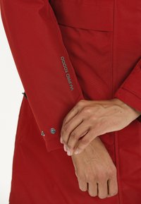 Punainen vedenpitävä takki, jossa on teksturoidut pinnat, W-PRO 10000 -merkintä, napsautettavat napit ja rannekkeiden säätimet.