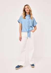 Light blue, short-sleeve denim shirt with a button-down front and tie detail at the waist, paired with white wide-leg pants and blue espadrilles.