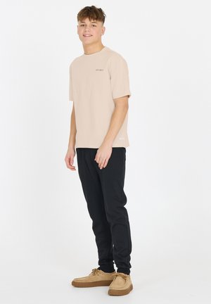 T-shirt beige clair à manches courtes, col ras du cou, avec un logo subtil, associé à un jogging noir et des chaussures à lacets beige sur un fond blanc.