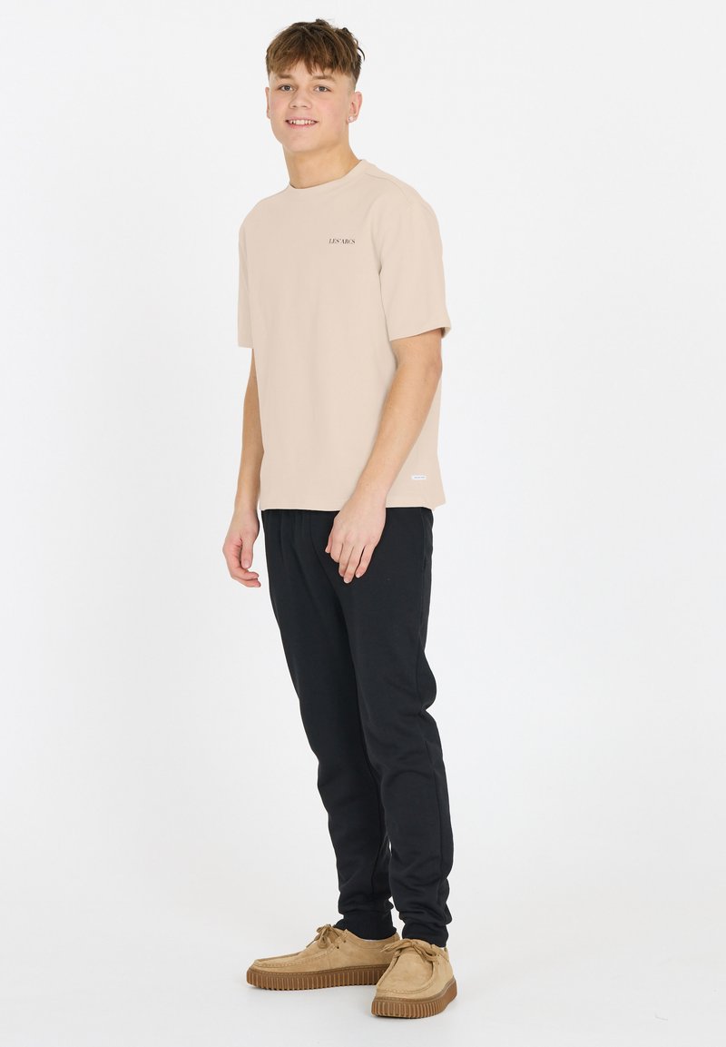 Lys beige t-shirt med korte ærmer, rund hals, og subtilt logo, parret med sorte sweatpants og lysebrune snørebåndssko på en hvid baggrund.