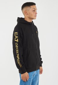 Felpa con zip nera realizzata in tessuto morbido. Presenta loghi dorati stampati sul petto e sulla manica. Include un cappuccio con cordino e tasche frontali.