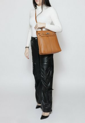 Donna che indossa un dolcevita bianco e pantaloni in pelle nera che tiene una borsa a tracolla in pelle marrone con fibbia argentata.