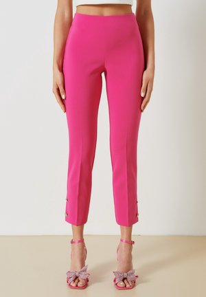 Kobieta ubrana w jasnoróżowe spodnie z wysokim stanem sięgające kostek z guzikami, zestawione z pasującymi różowymi szpilkami ozdobionymi dużymi kokardami.