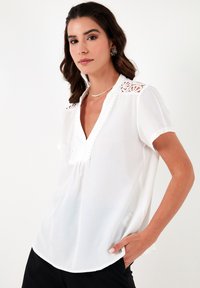 White short-sleeve blouse with a V-neck and lace detailing on shoulders. Soft, lightweight fabric with curved hem. Worn with black pants.