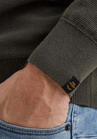 Oljčno zelena pletena manšeta puloverja z rebrastim potiskom in črno etiketo z napisom "PME Legend" v rumeni barvi. Roka, ki počiva na svetlo modri denim površini.