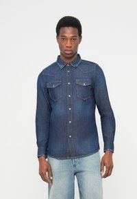 Tumši zils džinsa krekls ar izteiktu apkakli, divām krūšu kabatām ar aizsargām un pogu aizdari. Garas piedurknes ar pogām aprocēs.