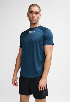 Man i marinblå Hummel sportst-shirt och svarta shorts står mot en enfärgad bakgrund, vänd framåt med neutralt uttryck.