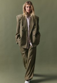 Olīvzaļa pārāk liela izmēra uzvalks ar dubultu krūšu mēteļa un platām biksēm, apvienots ar gaiši rozā svītrainu kreklu zem tā.