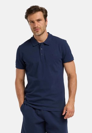 Uomo che indossa una polo blu navy a maniche corte e shorts blu navy abbinati, in piedi con una mano in tasca davanti uno sfondo uniforme.