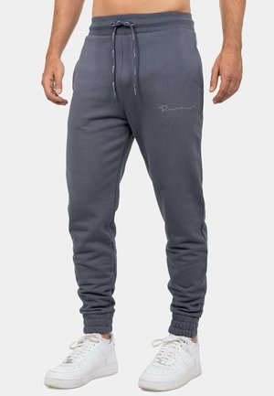 Hombre con pantalones jogger gris oscuro con cintura de cordón y puños elásticos, combinados con zapatillas deportivas blancas con cordones, de pie frente a un fondo liso.