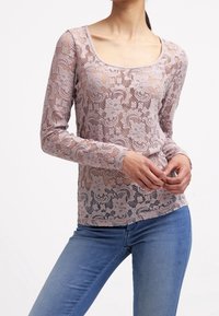 Blusa de encaje de mangas largas en rosa claro con patrón floral y escote redondeado. Tela semitranslúcida; combinada con jeans azules.