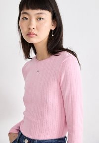 Lyserød langærmet top med ribbet tekstur og dekorative mønstre, med en rund halsudskæring og logo detalje på brystet.