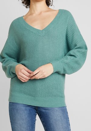 Femme portant un pull en maille turquoise à col en V avec des manches bouffantes et un jean bleu, debout, les mains jointes à la taille.