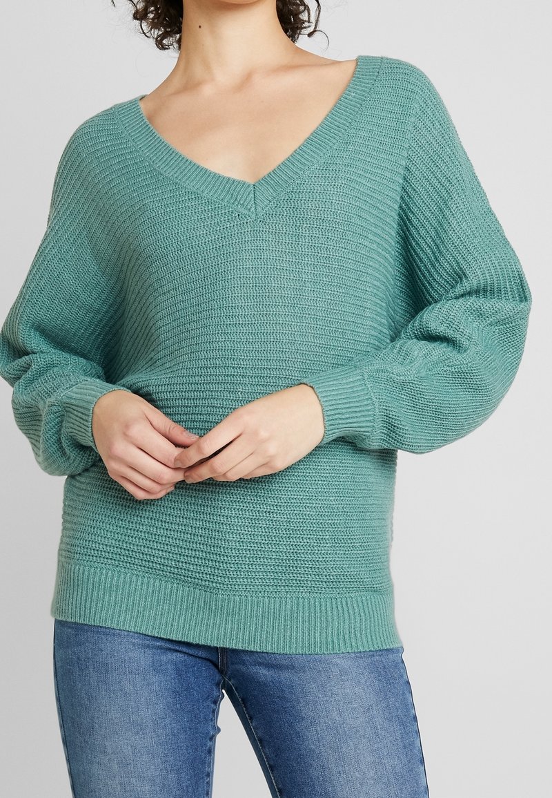 Femme portant un pull en maille turquoise à col en V avec des manches bouffantes et un jean bleu, debout, les mains jointes à la taille.