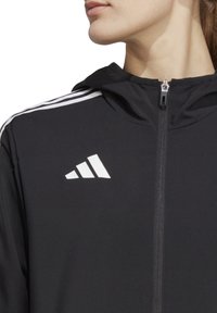 Giacca nera con zip e cappuccio, con logo Adidas bianco e tre strisce bianche sulle spalle. Tessuto dalla texture liscia.